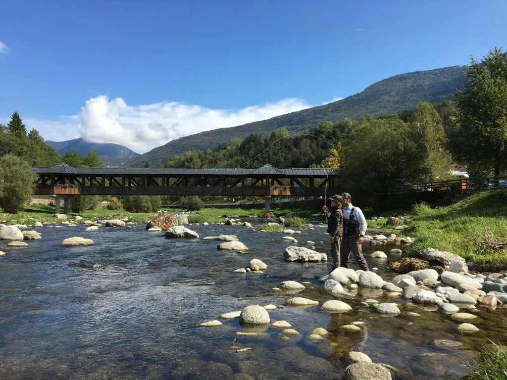 Pescatori a mosca in un fiume con ponte coperto e montagne sullo sfondo